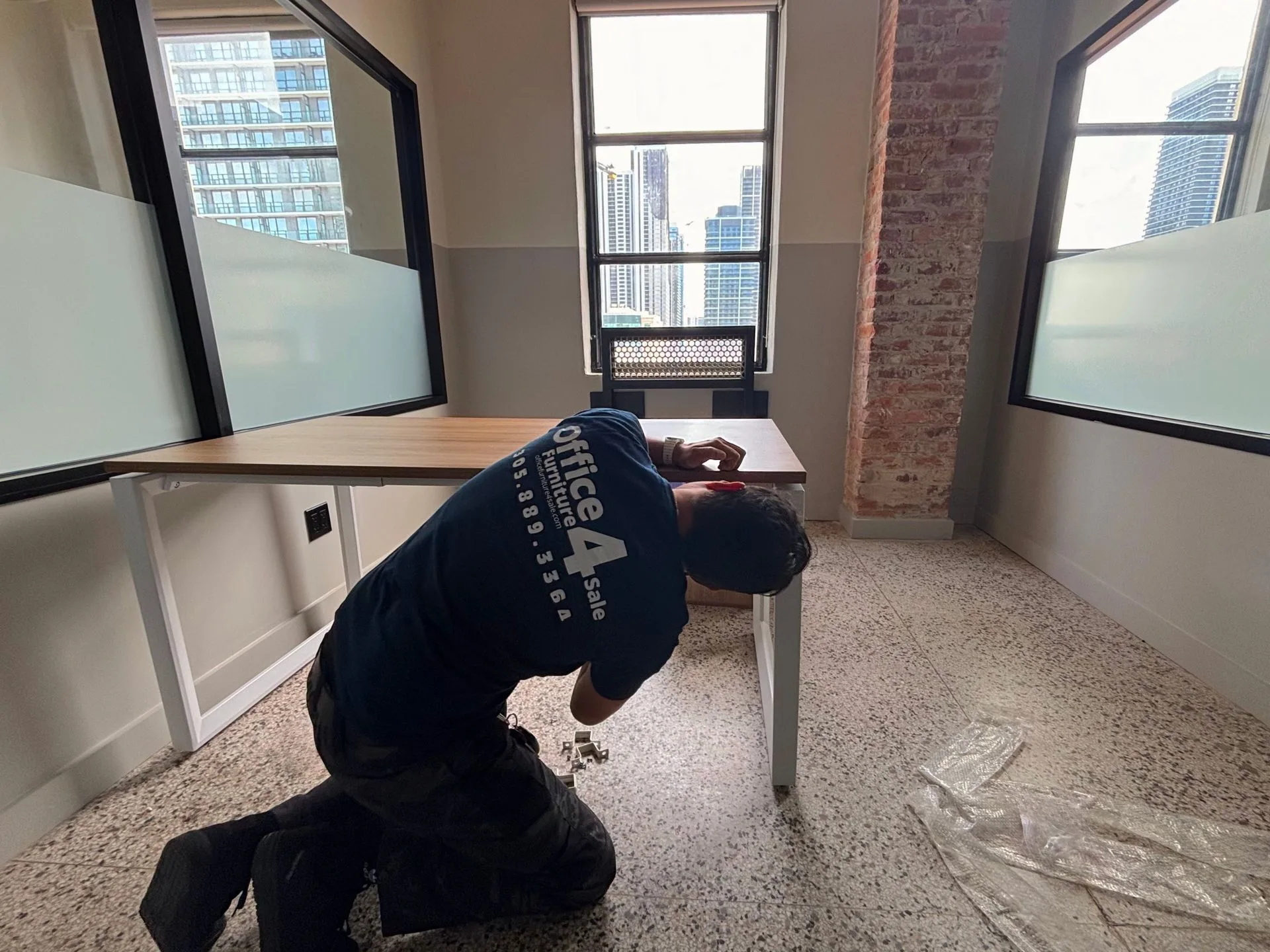 An OF4S installer assembling furniture in an office with a brick column and city-view windows