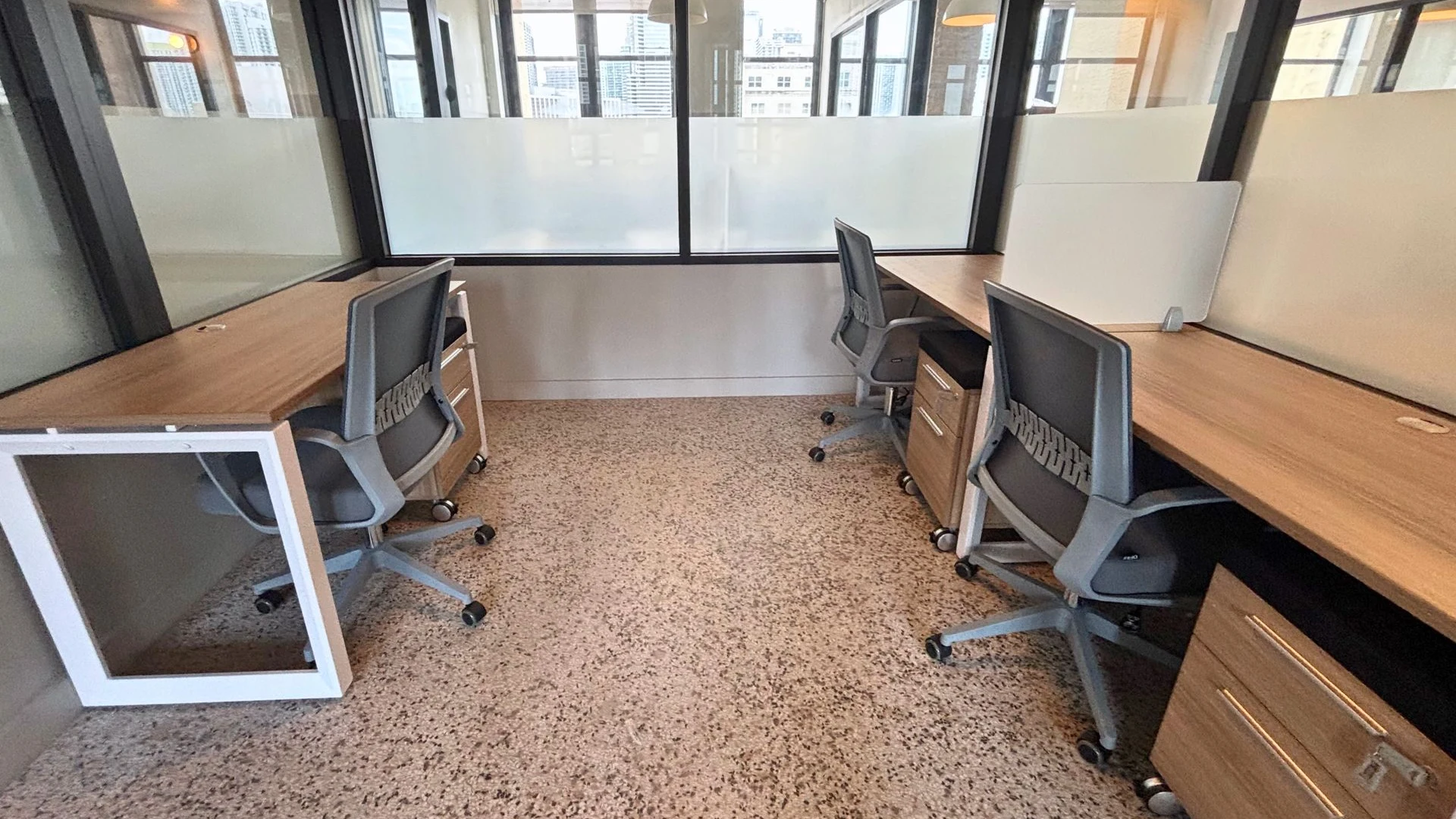 Two-person office with two facing wood desks, grey task chairs and a glass wall by the windows