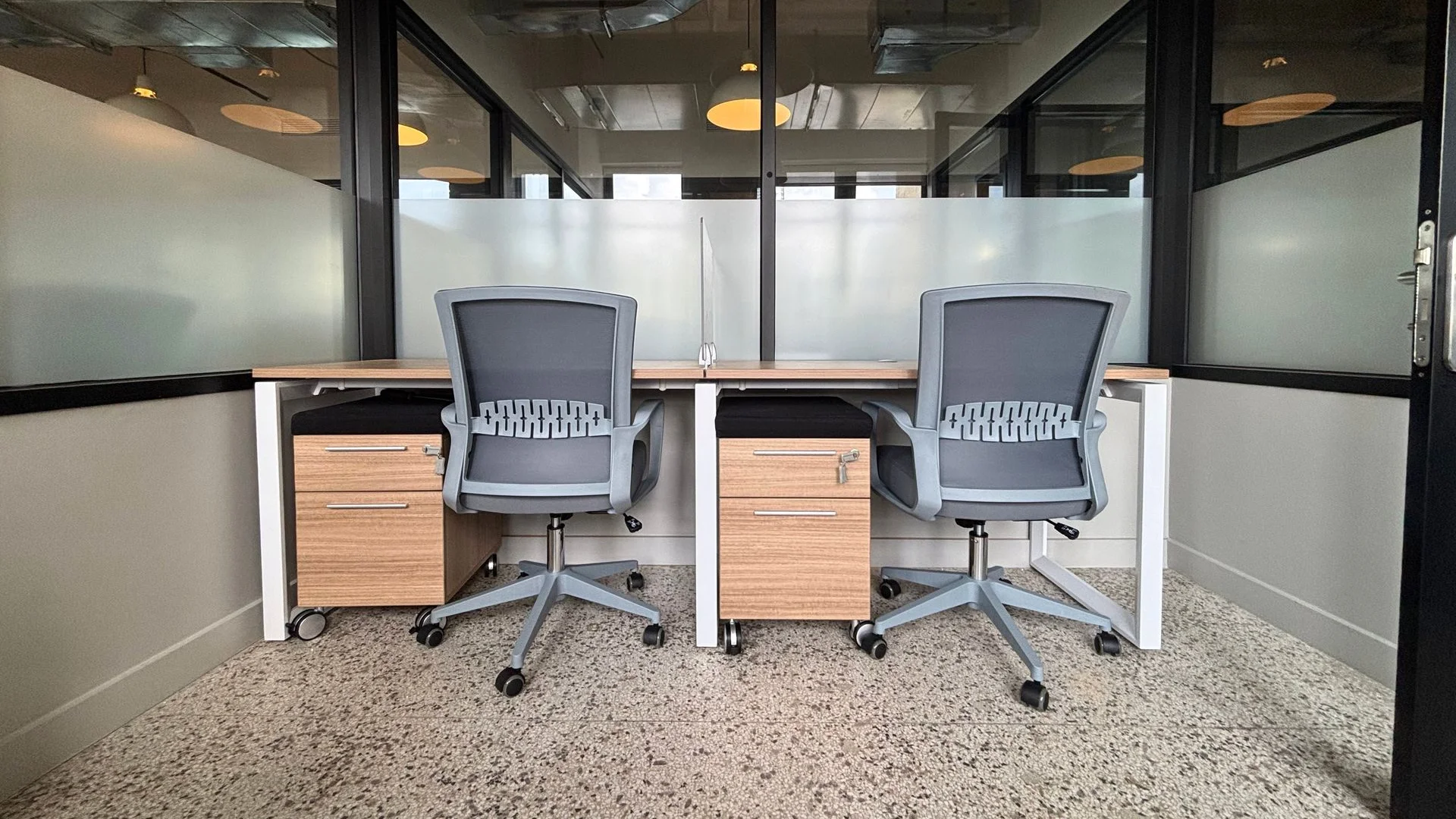 Two-person office with facing wood desks, grey task chairs and glass partitions