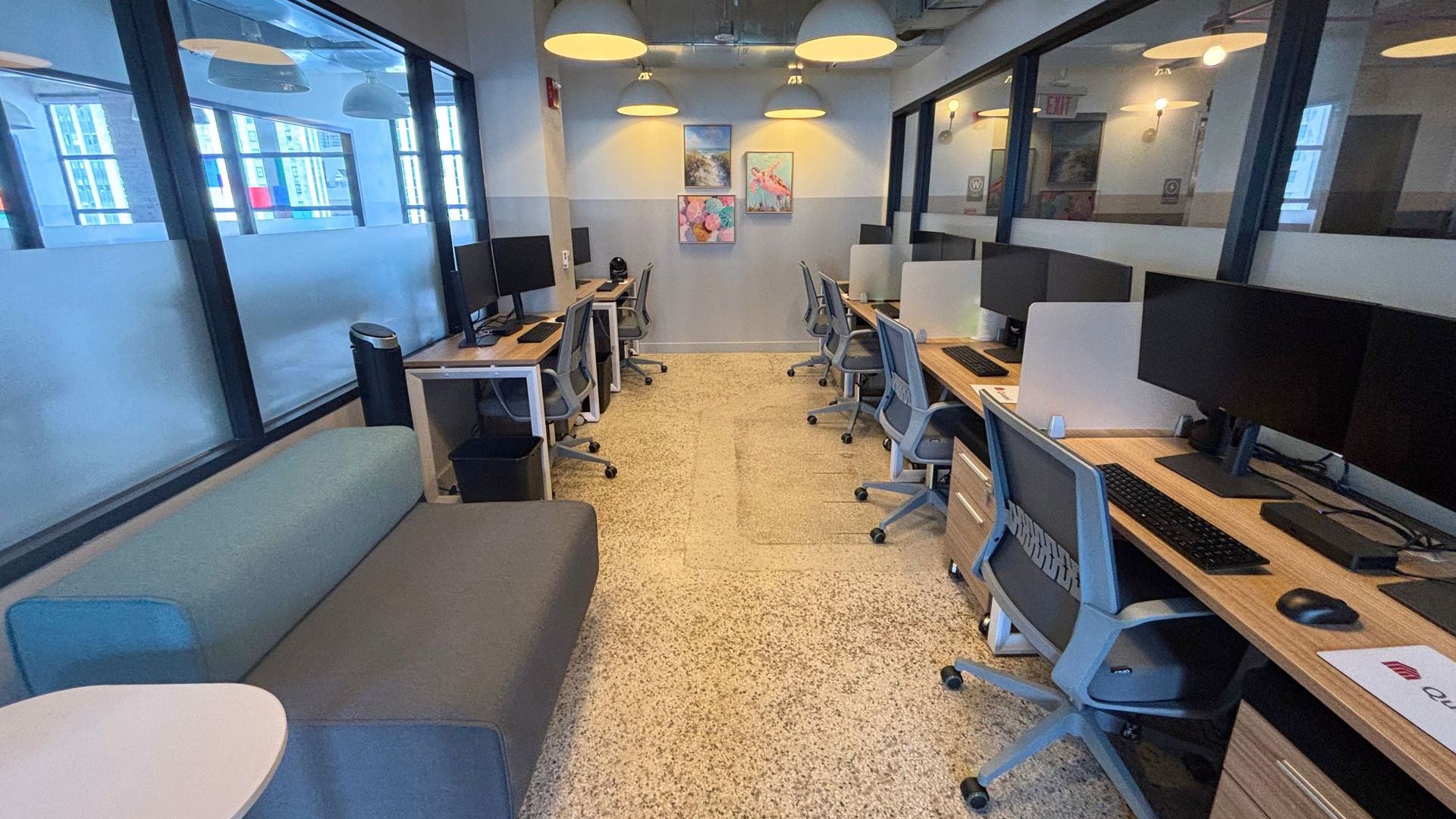 Open-plan workstation floor with a long run of wood bench desks, monitors and grey task chairs on terrazzo flooring