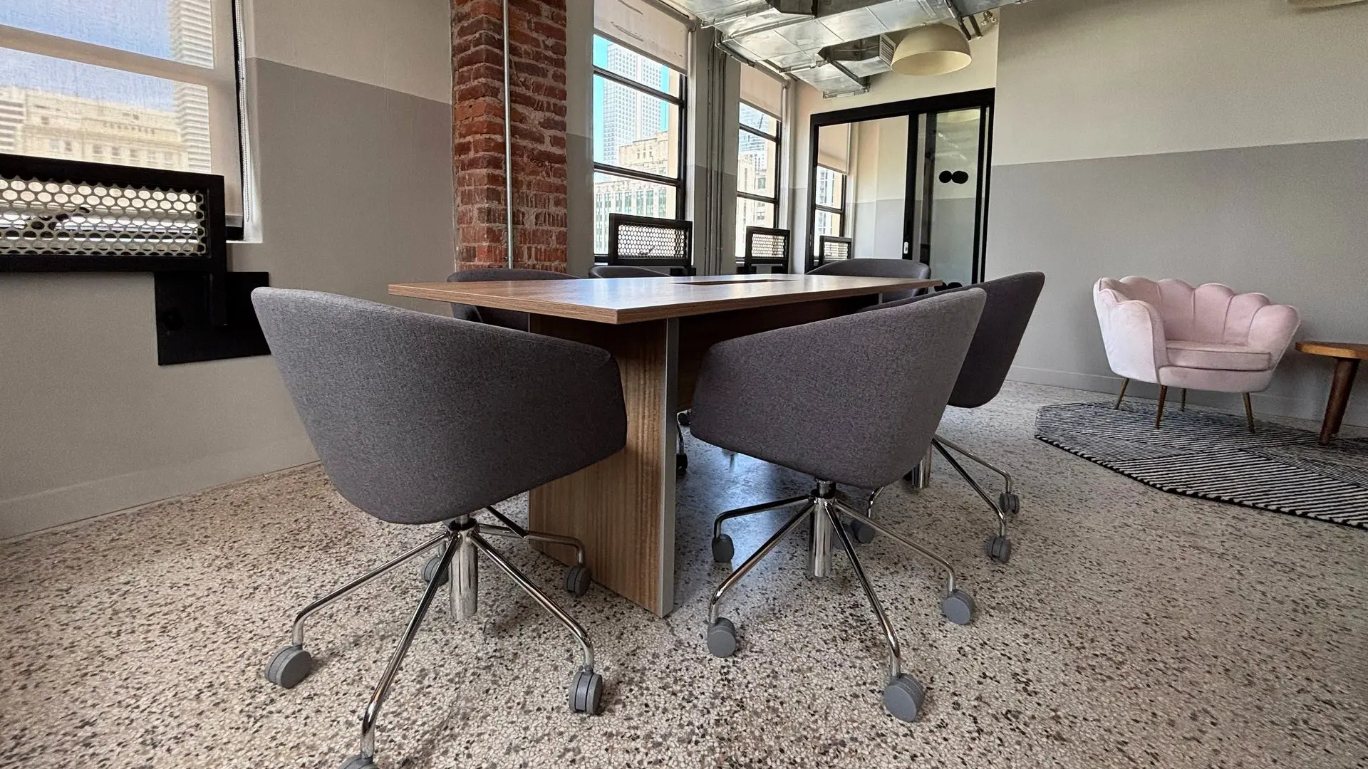 Meeting area with a round wood table, grey upholstered chairs and a blush accent chair beside a brick column