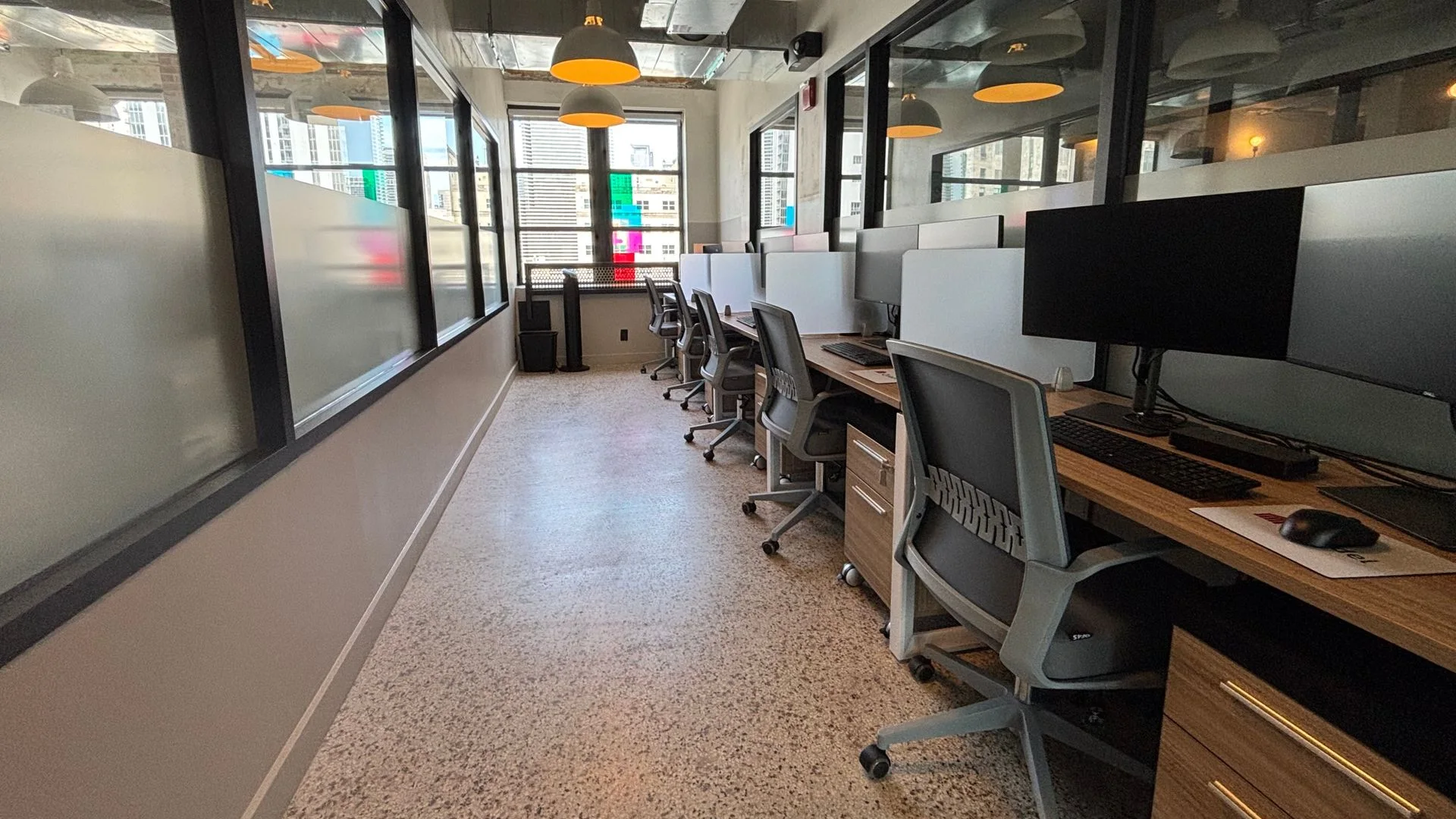 Open workstation floor with wood desks, monitors and grey task chairs under pendant lighting