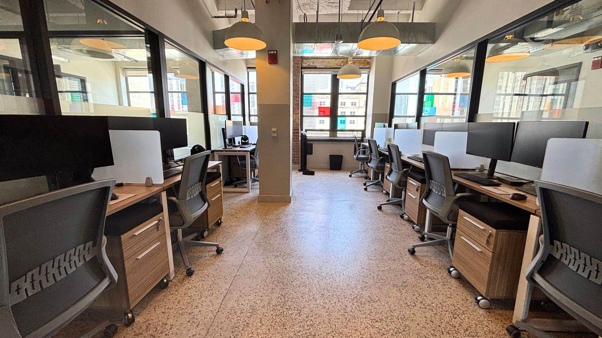 Long run of wood bench desks with monitors and grey task chairs beside the windows